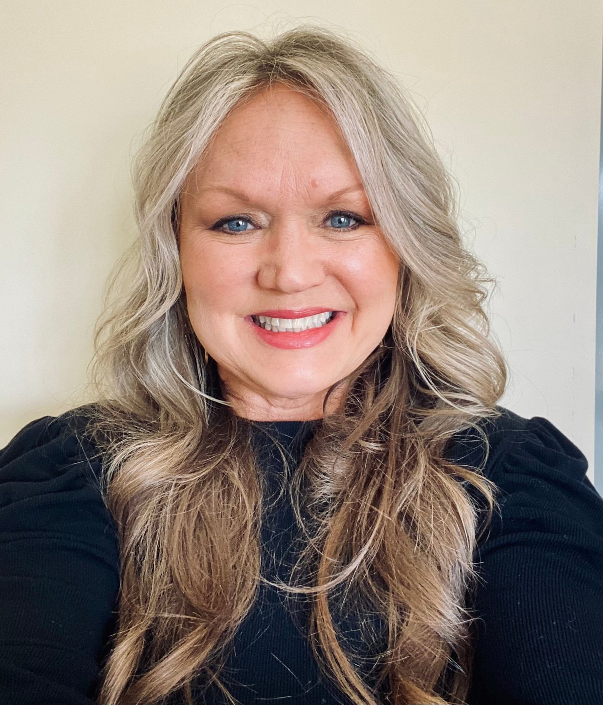Woman with long blonde wavy hair wearing a black top, smiling at the camera against a neutral beige background